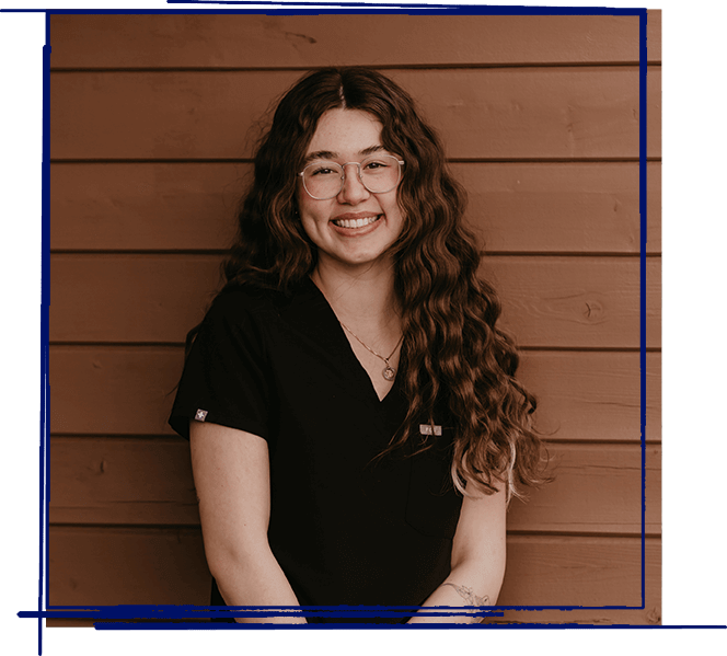 Smiling dental professional wearing glasses and black scrubs against a wooden background.