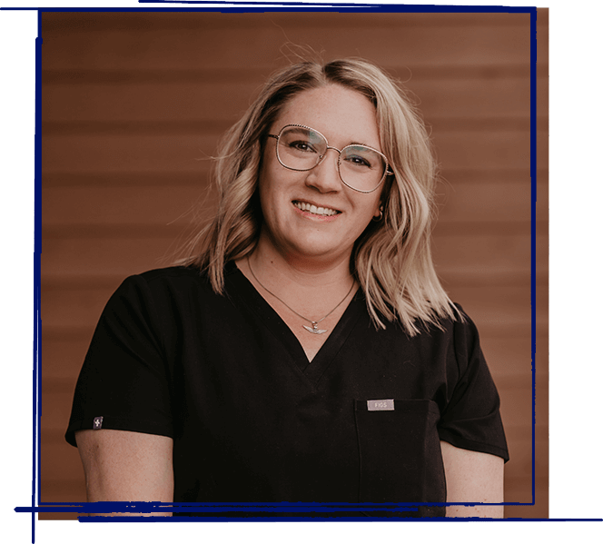 Woman wearing glasses and black scrubs smiling against brown wooden background.