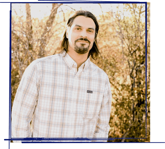 Man with long dark hair and goatee wearing plaid shirt outdoors with blurred trees background.