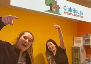 Two young women celebrate with raised arms in a bright orange dental office with Clubhouse Pediatric Dentistry signage.