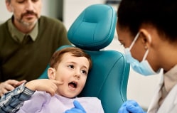 Young child in dental chair with open mouth during examination by pediatric dentist wearing mask.